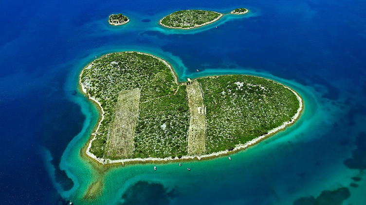 Aerial view of a heart-shaped island surrounded by turquoise waters, lush greenery covering the land, and a clear blue sky in the background.